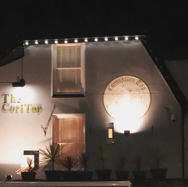 The front of the Coronation Tap at night, lit up with its round plaque