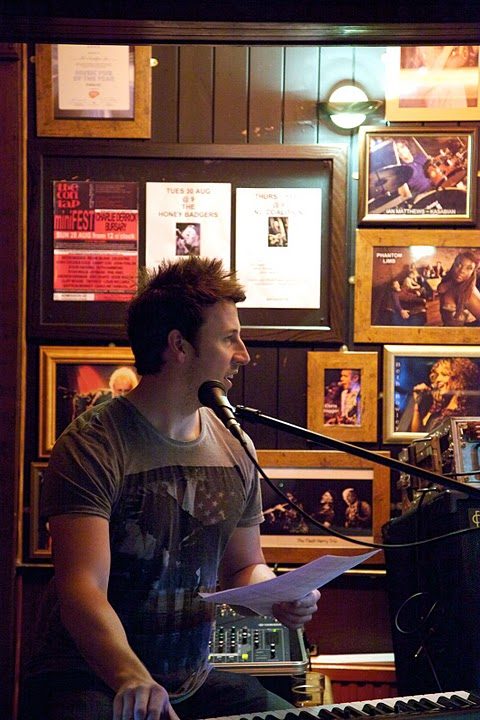 A musician playing keyboard and singing in front of the framed photos on the panelled wall